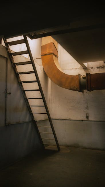 An industrial interior space featuring a black metal ladder positioned against a light-colored wall, leading to an upper level. Adjacent to the ladder, a large, curved, rust-colored metal duct runs from the ceiling downward, connected to a straight pipe segment. The floor appears to be concrete and is relatively clean, with minimal dust or debris visible. The lighting is dim, with natural light filtering in softly, highlighting the utilitarian environment. The scene illustrates the importance of surface management and inspection in areas requiring maintenance or cleaning, as emphasized by Kingston Carpet Cleaners in their guide on basement access issues for effective deep cleaning and sanitisation of such spaces.