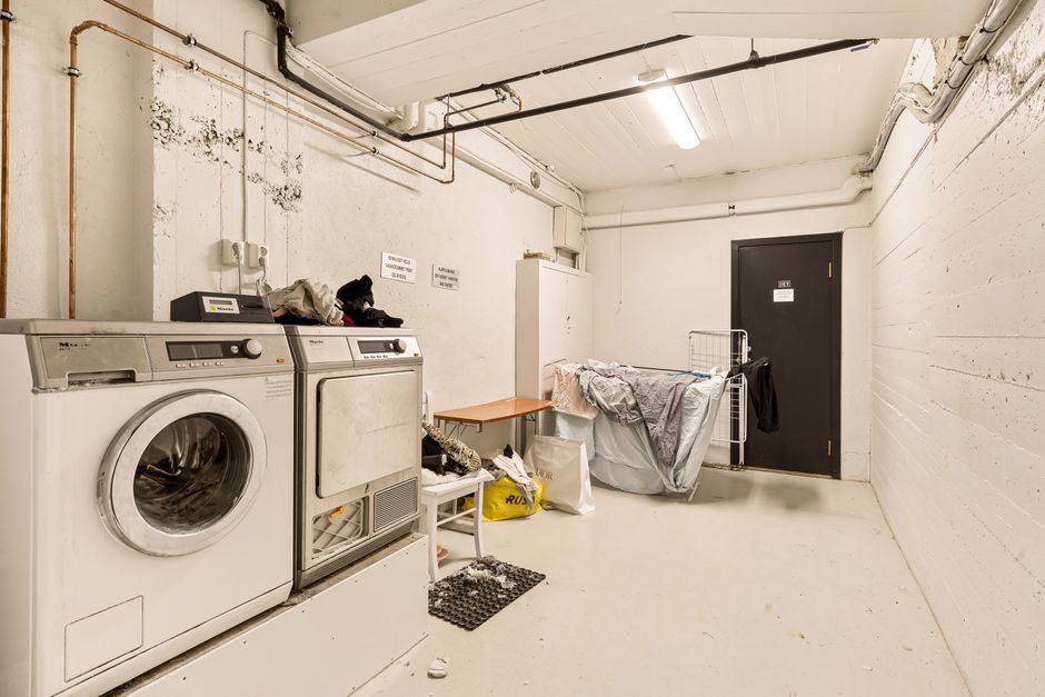 An industrial basement with concrete walls and ceiling, illuminated by a central long fluorescent light fixture, featuring a grey metal door with a small vertical window. The walls show a raw cement finish, with visible piping and an orange emergency alarm box attached to the left wall. The floor appears clean and smooth, indicating recent surface cleaning or maintenance, reflecting a well-maintained space suitable for access issues for basement cleaning in Kingston, as addressed by Kingston Carpet Cleaners.