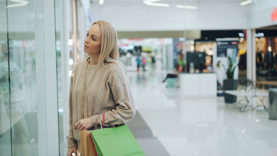 A young blonde woman with straight hair, dressed in a beige knitted sweater, is standing inside a shopping mall near a glass storefront, holding green and red shopping bags. The shopping mall features glossy tiled floors, modern furniture, and bright overhead lighting that reflects off the clean, polished surfaces. The background includes shops, seating areas, and a few shoppers walking or sitting. The overall scene emphasizes cleanliness, surface maintenance, and the importance of hygiene in retail environments. Kingston Carpet Cleaners, specializing in surface cleaning and sanitisation, highlights the significance of deep cleaning for retail spaces like Bentall Centre in Kingston as part of their professional cleaning services.