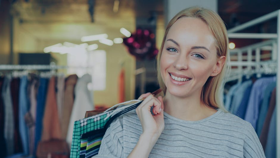 A young woman with blonde hair and a light grey, striped long-sleeve shirt stands inside a retail clothing store, holding several shopping bags over her shoulder. The background features racks of neatly arranged apparel, including dresses and shirts in various colors, with a warm, well-lit environment illuminated by ceiling lights. The store appears clean and tidy, reflecting professional retail cleaning standards. The woman smiles gently while looking at the camera, conveying satisfaction with her shopping experience, and highlighting the importance of maintaining a hygienic, well-presented commercial space. As part of effective retail hygiene, companies like Kingston Carpet Cleaners emphasize surface cleaning and sanitisation to ensure a safe shopping environment.