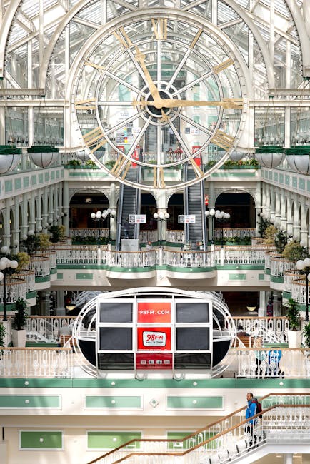 A young blonde woman with straight hair, dressed in a beige knitted sweater, is standing inside a shopping mall near a glass storefront, holding green and red shopping bags. The shopping mall features glossy tiled floors, modern furniture, and bright overhead lighting that reflects off the clean, polished surfaces. The background includes shops, seating areas, and a few shoppers walking or sitting. The overall scene emphasizes cleanliness, surface maintenance, and the importance of hygiene in retail environments. Kingston Carpet Cleaners, specializing in surface cleaning and sanitisation, highlights the significance of deep cleaning for retail spaces like Bentall Centre in Kingston as part of their professional cleaning services.