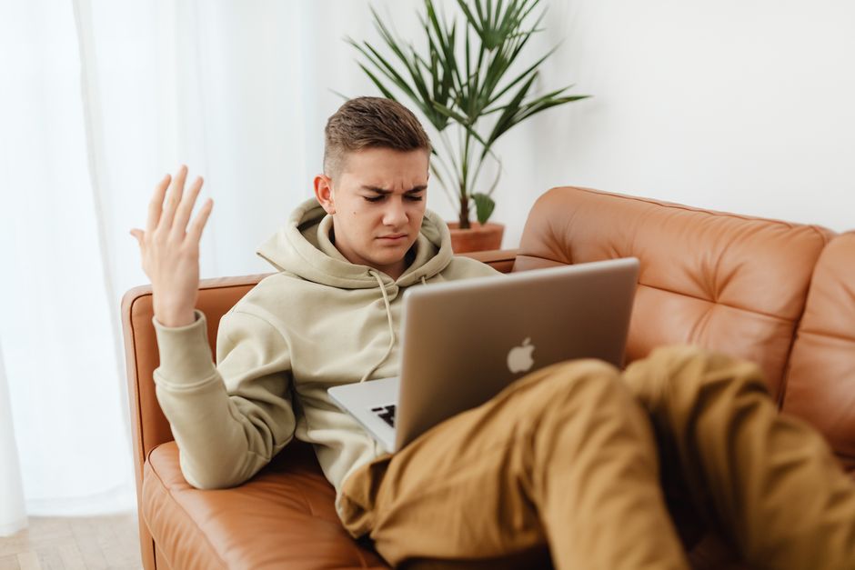 A young man with short brown hair is seated on a tan leather sofa in a bright living room, holding a laptop on his lap. He has a frustrated expression, with one hand raised and eyebrows furrowed, indicating confusion or concern. The room features neutral-colored walls, a potted plant with large green leaves positioned on a wooden side table behind him, and sheer white curtains allowing natural light to fill the space. The surface around him appears clean and well-maintained, with no visible dust or clutter. This setting illustrates a domestic environment where Kingston Carpet Cleaners offers professional cleaning services to ensure surface hygiene and maintain a fresh, sanitized home.