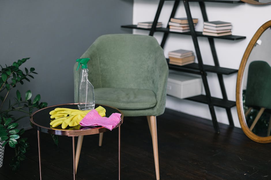 A modern living room features a light green upholstered armchair positioned near a dark wooden floor. On a small, round black side table with a metallic rose gold rim, there are yellow rubber cleaning gloves, a pink cloth, and a transparent spray bottle with a green trigger, indicating cleaning supplies. To the left, a potted green houseplant adds a touch of nature, while in the background, a black ladder-style bookshelf holds neatly stacked books. An oval mirror with a wooden frame rests against the wall, reflecting part of the room. The room appears tidy and well-lit, with natural light accentuating the cleanliness of the surfaces, illustrating the importance of surface cleaning and maintenance promoted by Kingston Carpet Cleaners.