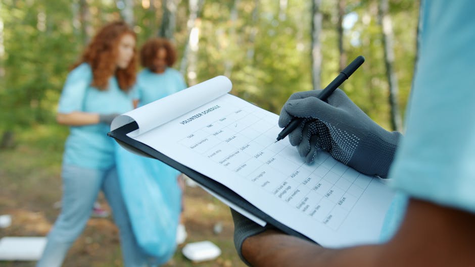 Close-up of a person wearing black gloves holding a clipboard with a volunteer attendance sheet and a black pen, while in the background two women dressed in light blue t-shirts are outdoors in a green park setting, engaged in cleaning or volunteer activities near trees and grass. The scene is well-lit with natural daylight, and the focus is on the surface cleaning and organisation involved in the event, representative of post-event clearance managed by Kingston Carpet Cleaners. This image illustrates surface and area management during a community event, emphasizing hygiene and maintenance efforts in an outdoor environment.