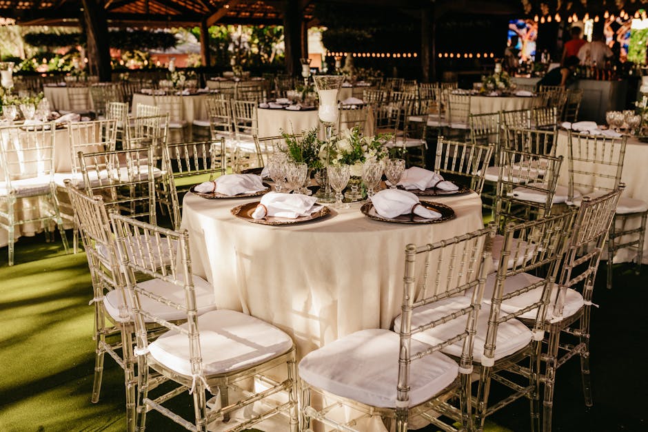 A decorated event space with round tables covered in cream tablecloths and surrounded by silver chiavari chairs with white cushions. Each table is set with decorative glassware, including water goblets and wine glasses, and white napkins neatly folded on dark, round chargers. A tall glass candle holder with a white candle and floral arrangements with greenery serve as centerpieces on each table. The venue has a wooden gazebo structure with string lights hanging from the ceiling, creating a warm, inviting atmosphere. Natural light filters in from the open sides, illuminating the polished floors and highlighting the clean, well-maintained surfaces. The setting appears ready for a formal gathering or celebration, with attention to detail in both decoration and cleanliness, reflecting the professional cleaning standards of Kingston Carpet Cleaners in maintaining hygienic, visually appealing event environments.