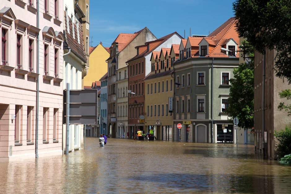 A flooded urban street scene with several multi-story buildings featuring colorful facades in shades of pink, yellow, orange, green, and beige, and red-tiled roofs. The street is submerged under a layer of muddy water, reaching the base of the buildings and partially covering the sidewalks. Few people are visible walking through the water, and some are wearing high-visibility clothing, possibly emergency responders. The sky is clear with a few clouds, and sunlight illuminates the scene, highlighting the wet surfaces. The image showcases the impact of urban flooding, emphasizing the importance of prompt emergency flood cleanup and surface sanitisation, as offered by Kingston Carpet Cleaners.