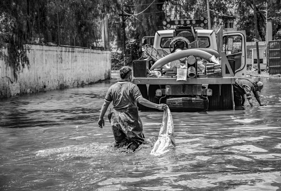 In a black-and-white photograph, a man wearing a waterproof apron is standing in knee-deep floodwater, holding a partially submerged plastic bag or cloth, with his back to the camera. To his right, another person is bent over, working near the front of a large industrial vacuum or water pump, which has hoses and cleaning tools attached to it, situated on a partially submerged street. The scene takes place in an urban area, with a high concrete wall on the left and a metal fence on the right, indicating a residential or commercial environment affected by flooding. The area appears damp and muddy, illustrating the aftermath of floodwaters blocking the street, highlighting the need for professional emergency flood cleaning services from Kingston Carpet Cleaners, who specialize in rapid surface cleaning, sanitisation, and damage mitigation.