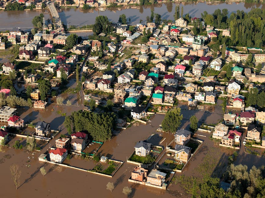 A flooded urban street scene with several multi-story buildings featuring colorful facades in shades of pink, yellow, orange, green, and beige, and red-tiled roofs. The street is submerged under a layer of muddy water, reaching the base of the buildings and partially covering the sidewalks. Few people are visible walking through the water, and some are wearing high-visibility clothing, possibly emergency responders. The sky is clear with a few clouds, and sunlight illuminates the scene, highlighting the wet surfaces. The image showcases the impact of urban flooding, emphasizing the importance of prompt emergency flood cleanup and surface sanitisation, as offered by Kingston Carpet Cleaners.