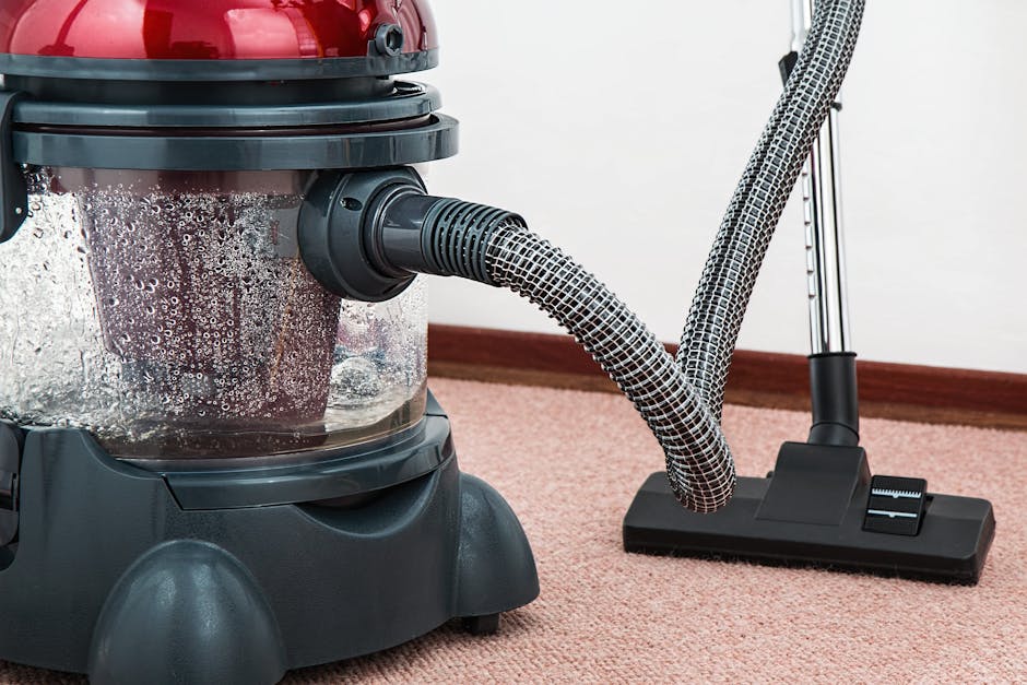 Close-up of a commercial carpet cleaning vacuum with a transparent water tank filled with soapy water, connected to a flexible hose and a cleaning head resting on a pinkish-beige carpeted floor in a room with white walls. The vacuum is in use as part of a surface cleaning or deep cleaning process, with visible foam bubbles indicating recent operation, and is positioned near a wooden skirting board. The setting suggests hygienic and effective carpet maintenance by Kingston Carpet Cleaners, emphasizing professional cleaning services in a domestic or commercial environment.