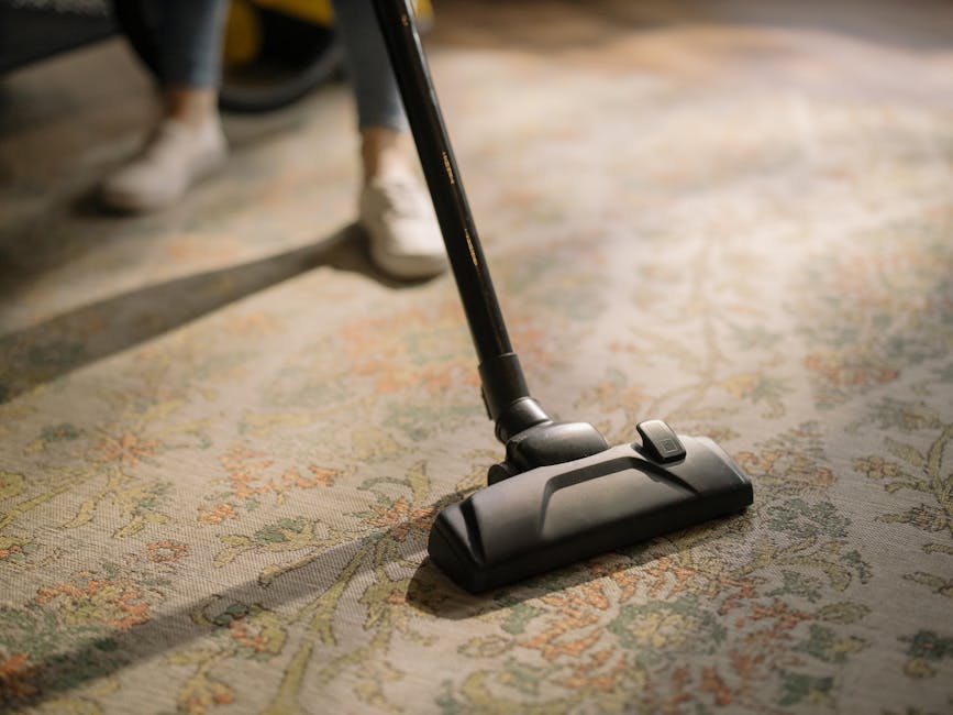 Close-up of a commercial carpet cleaning vacuum with a transparent water tank filled with soapy water, connected to a flexible hose and a cleaning head resting on a pinkish-beige carpeted floor in a room with white walls. The vacuum is in use as part of a surface cleaning or deep cleaning process, with visible foam bubbles indicating recent operation, and is positioned near a wooden skirting board. The setting suggests hygienic and effective carpet maintenance by Kingston Carpet Cleaners, emphasizing professional cleaning services in a domestic or commercial environment.