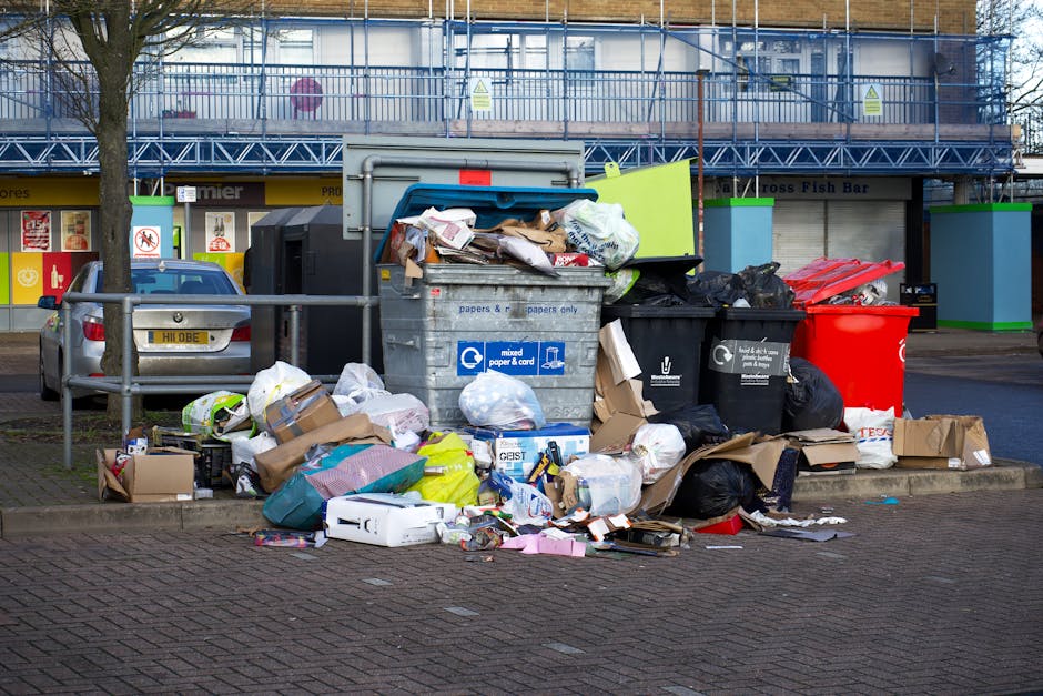 A black waste bin labeled 'St. John's' positioned on a residential sidewalk at night, filled with various domestic waste including cardboard boxes and paper. The bin is situated next to a curb under f