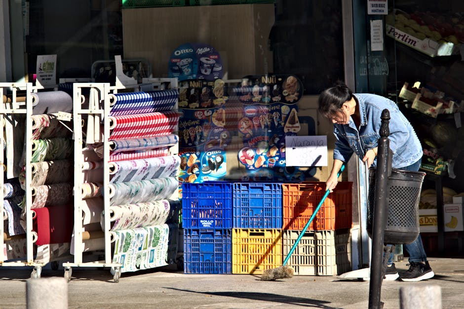 A woman wearing a denim jacket and black sneakers is seen sweeping the sidewalk outside a store that sells rolls of patterned wallpaper and ice cream products displayed behind glass. She is using a light blue-handled broom to clean the pavement, which appears to be in good, clean condition with minimal debris. The store window features colorful ice cream packaging and Oreo-themed products, with a white sign displayed in the window. The scene is illuminated by natural daylight, highlighting the storefront's exterior surfaces and the woman’s focused cleaning activity, illustrating commercial cleaning practices relevant to maintaining hygiene and cleanliness for shop fronts, as promoted by Kingston Carpet Cleaners on their Kingston Station advice page.