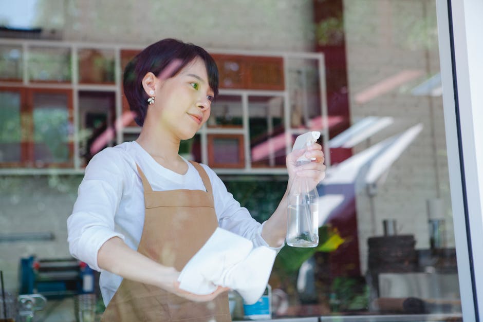 A woman with short dark hair and pearl earrings is depicted cleaning a large glass window, holding a spray bottle in her right hand and a white cloth in her left. She is wearing a white long-sleeved shirt under a light brown apron, emphasizing her role in surface cleaning or sanitisation. The background shows a bright, modern interior with a wooden bookshelf filled with books, potted plants, and a countertop, illuminated by natural light coming through the window. The glass appears clean and shiny, reflecting the indoor environment and outdoor scenery, highlighting the thoroughness of her cleaning process suitable for storefront or commercial window maintenance, aligning with Kingston Carpet Cleaners’ expertise in professional cleaning services.