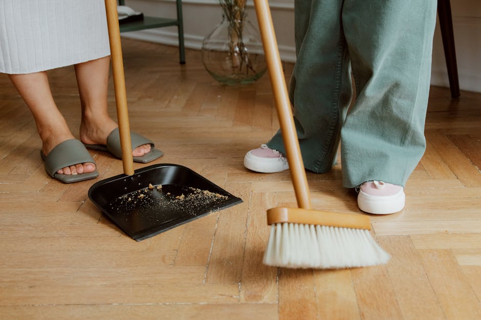 Close-up view of two individuals sweeping a light-colored wooden floor in a residential setting. One person, wearing gray slippers and a white dress, holds a black dustpan with some crumbs inside, whi