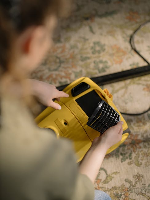 A person holds a yellow portable fan heater or dehumidifier over a patterned carpet in a room with soft lighting, suggesting a focus on surface cleaning and drying as part of deep cleaning or sanitisation. The carpet features a traditional floral design with muted colors, and the scene is captured from above, emphasizing the equipment being used for thorough cleaning. Visible cords indicate electrical power, and the image complements the services offered by Kingston Carpet Cleaners, highlighting their expertise in domestic cleaning and fast drying solutions to address delays and hidden charges as mentioned in the page title.