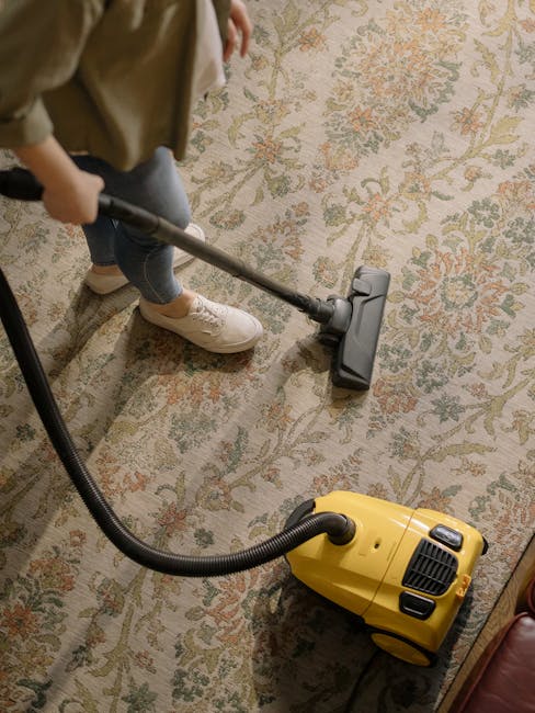 A person holds a yellow portable fan heater or dehumidifier over a patterned carpet in a room with soft lighting, suggesting a focus on surface cleaning and drying as part of deep cleaning or sanitisation. The carpet features a traditional floral design with muted colors, and the scene is captured from above, emphasizing the equipment being used for thorough cleaning. Visible cords indicate electrical power, and the image complements the services offered by Kingston Carpet Cleaners, highlighting their expertise in domestic cleaning and fast drying solutions to address delays and hidden charges as mentioned in the page title.